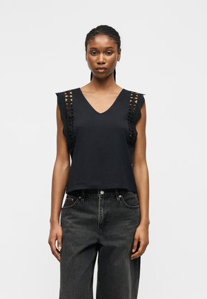 Ung kvinde iført en sort ærmeløs top med hæklet blonde detaljer og højtaljede, udvaskede sorte jeans, står foran en ensfarvet baggrund.