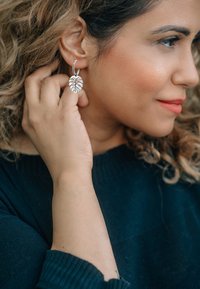 Orecchini a cerchio in argento con un design dettagliato a foglia, che mettono in mostra motivi a intaglio e una finitura lucida, indossati da una persona con capelli ricci.