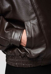 Mano en el bolsillo de una chaqueta de cuero marrón oscuro con textura, con cintura y puño elásticos sobre fondo claro.