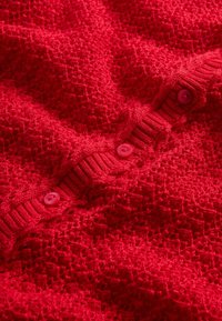 Sarkans adīts kardigans ar teksturētu rakstu, ribām aproču malās un rozā apaļām pogām pie priekškadara slēgšanas.