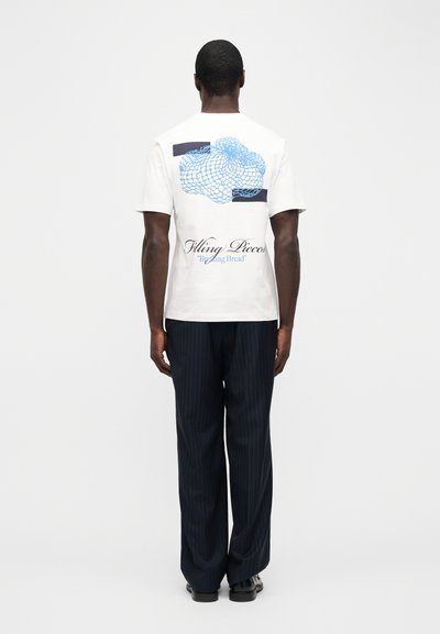 Άνδρας που φοράει λευκό μπλουζάκι με μπλε αφηρημένο σχέδιο και το κείμενο "Filling Pieces 'Breaking Bread'" στην πλάτη, σε συνδυασμό με σκούρο παντελόνι με λεπτές κάθετες ρίγες.