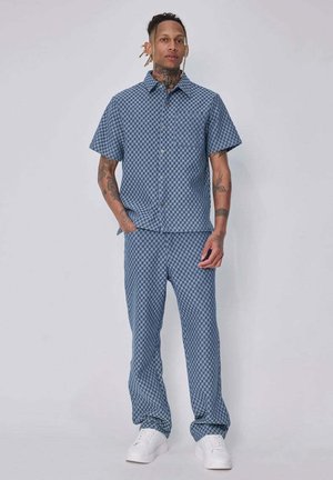 Homme avec des tatouages et des dreadlocks porte une chemise à manches courtes à carreaux bleue et un pantalon assorti avec des baskets blanches, debout devant un fond gris.