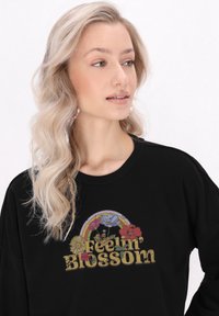 Fekete pulóver, színes virág- és szivárvány grafikával, amelyen a "Feelin' Blossom" felirat található, textúrált, vastag betűtípussal.