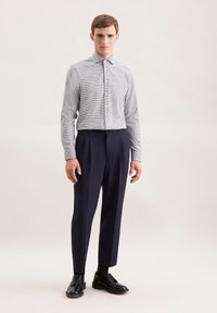 Uomo che indossa una camicia button-up a quadri bianchi e neri, pantaloni plissettati blu navy, calze nere e scarpe eleganti nere, in piedi di fronte a uno sfondo neutro.