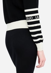 Maglione nero lavorato a maglia con strisce bianche, orlo a coste e dettagli in perle sulle maniche. Presenta un logo in testo a contrasto.