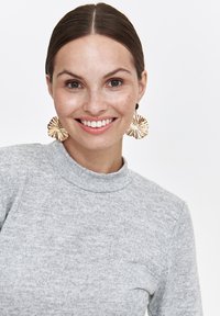 Lächelnde Frau mit braunem, zurückgezogenem Haar, die große goldene Blumenohringe und einen hellgrauen Strickpullover mit hohem Kragen trägt.