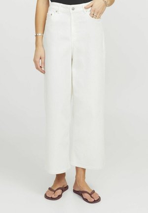Pantalones de mezclilla anchos de color blanco con una cintura alta, cuentan con bolsillos frontales y una cremallera. Combinados con chanclas marrones.