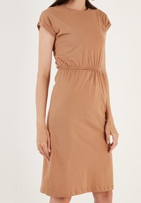 Lyhythihainen ruskea mekko, joka on valmistettu pehmeästä kankaasta ja jossa on joustava vyötärö. Mekko ulottuu juuri polven ylle, ja siinä on pyöreä kaula-aukko sekä käännetyt hihat.