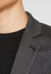 Gros plan d'une personne portant un blazer à carreaux noir et gris texturé avec une petite épingle de revers en forme de feuille sombre et un pull en maille noire.