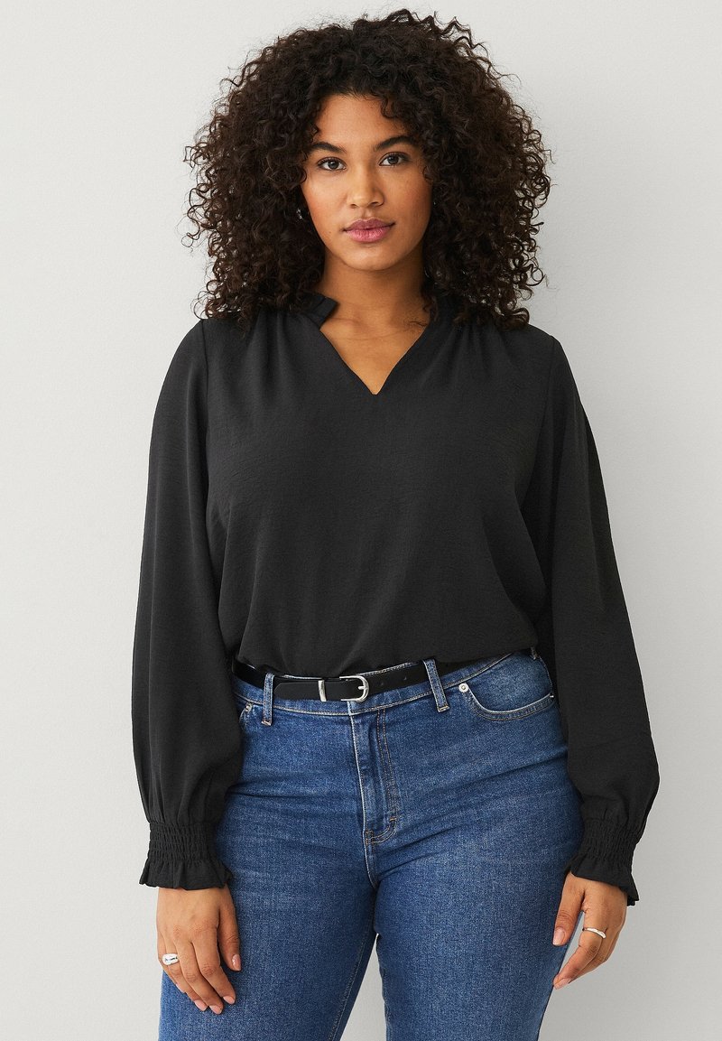 Blusa negra de escote en V con mangas largas y recogidas, llevada sobre unos vaqueros azules. La tela tiene una textura suave y un corte relajado.