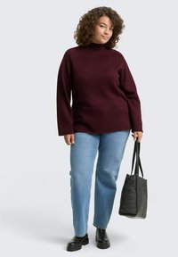 Pull col roulé bordeaux à manches longues, associé à un jean bleu clair et des bottines noires, tenant un sac fourre-tout noir.