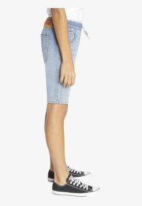 Shorts de mezclilla azul claro con cintura elástica, bolsillos laterales y una etiqueta de cuero en la parte posterior. Combinados con zapatillas de lona negras.