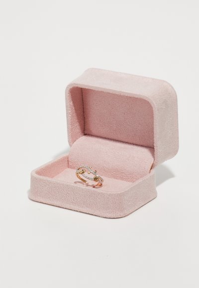 Ružová zamatová krabička na prstene s mäkkou textúrou, obsahujúca zlatý prsteň so sklenenými kameňmi, tvarovaný ako previazané vetvičky.