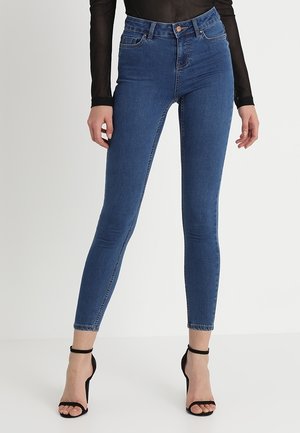 Niebieskie obcisłe jeansy z denimu noszone z czarnym przejrzystym topem z długim rękawem oraz czarnymi sandałami na wysokim obcasie z paskiem wokół kostki, stojące na białym tle.