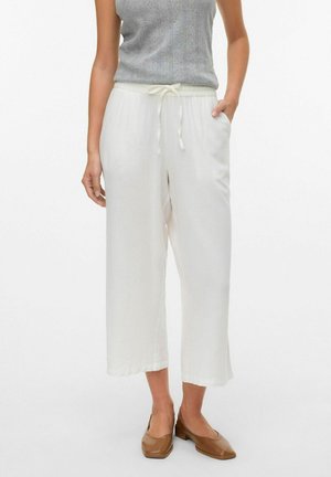 Femme portant un pantalon blanc large coupé aux chevilles avec une taille à cordon, un haut en tricot gris sans manches et des chaussures marron à petit talon, debout sur fond blanc.
