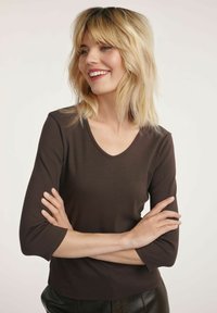 Blondynka z włosami do ramion uśmiecha się, nosząc ciemno-brązową bluzkę z rękawem 3/4 i czarne spodnie, z założonymi na siebie rękami.