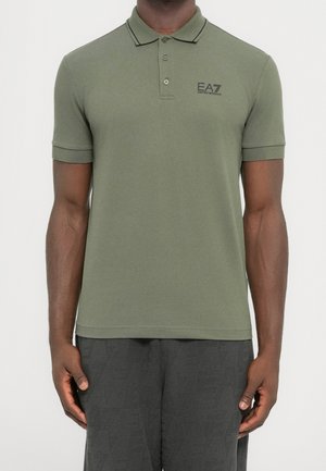 Man draagt een olijfgroen poloshirt met korte mouwen met het EA7 Emporio Armani-logo op de linkerborst, gecombineerd met een donkere gestructureerde broek.
