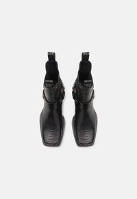 Paire de bottines noires avec texture peau de serpent, bouts carrés, sangles latérales à boucle et languettes à l'arrière sur un fond uni.