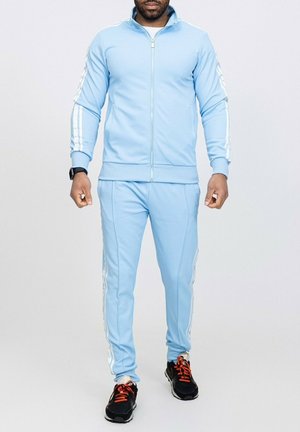 Survêtement bleu clair en tissu lisse, avec des rayures blanches le long des manches et sur les côtés, accompagné de chaussures de sport noires.