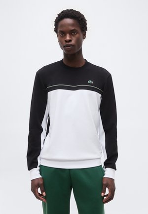 Mann trägt ein schwarz-weißes Langarm-Sweatshirt mit einem kleinen grünen Logo auf der Brust und grüne Hosen, steht vor einem schlichten Hintergrund.