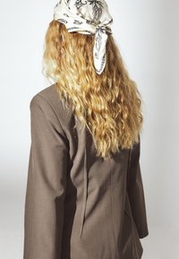 Blazer marrone con un design strutturato, caratterizzato da un collo alto e maniche lunghe. Lunghi capelli biondi e mossi sono parzialmente coperti da una sciarpa bianca con motivi neri.