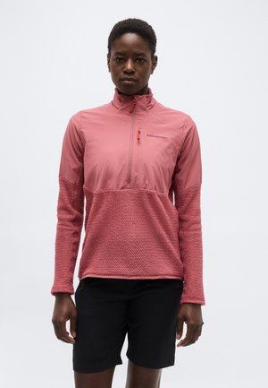 Mujer con chaqueta rosa texturizada con media cremallera y shorts negros, de pie frente a un fondo blanco liso.