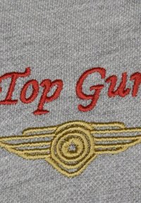 Szary materiał z czerwonym haftowanym napisem "Top Gun" nad żółtym, okrągłym emblematem z skrzydłami, ukazujący szczegółowe przeszycia.