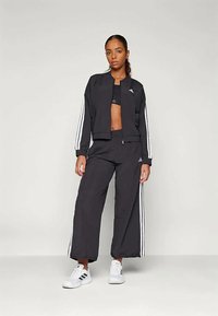 Fekete sportos melegítő szett, amely tartalmaz egy rövidített dzsekit és széles szárú nadrágot, oldalán fehér csíkkal, fehér sneaker cipőkkel párosítva.