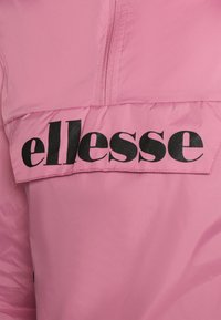 Rosa vindjacka med delvis dragkedja och en stor svart "ellesse" logotyp framtill. Slät, lätt material med matt yta.