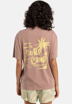 Persoon met krullend haar, gekleed in een mauve T-shirt met gele "Surf Camp"-opdruk en palmboomdesign, gecombineerd met groen gebloemde short, van achteren gezien.