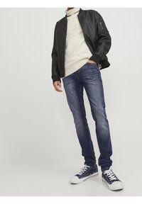 Černá bomber bunda, krémový rolák, slim-fit modré džíny a námořnické tenisky s bílými tkaničkami, na světle šedém pozadí.