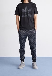 Man står med händerna i fickorna iförd svart Adidas t-shirt, mönstrade svarta Adidas träningsoverallsbyxor och grå Adidas sneakers på en blå golvyta.