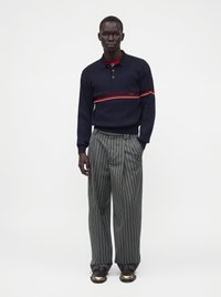 Homme portant un pull bleu marine à rayures rouges, un pantalon ample gris à fines rayures et des chaussures noires avec des bouts dorés, debout devant un fond blanc.