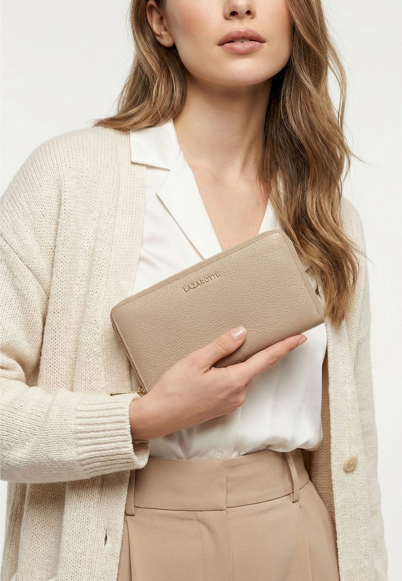 Mujer con cárdigan y pantalones beige sostiene una cartera de cuero beige con cremallera y etiqueta Lazarotti contra su pecho.
