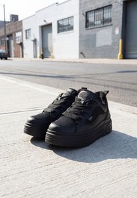 Baskets plateformes noires avec semelles épaisses et lacets, posées sur un trottoir urbain ensoleillé près de bâtiments industriels.