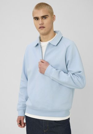 Jeune homme aux cheveux blonds courts portant un sweat-shirt bleu clair à fermeture éclair quart et un jean foncé, ajustant la fermeture éclair d'une main.