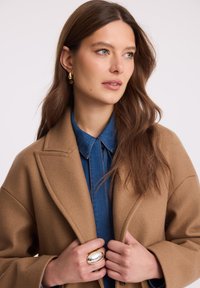 Kobieta z długimi brązowymi włosami, nosząca złote kolczyki, duży srebrny i złoty pierścień, niebieską koszulę oraz camelowy płaszcz, patrząca w bok.