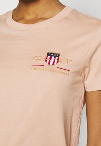 Camiseta de algodón melocotón con mangas cortas, cuello redondo y un logotipo bordado con diseño de escudo en tonos rojo, azul y dorado.