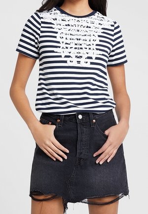 Kvinde iført navyblå og hvide stribede top med korte ærmer med hvide blomstrede blonder detaljer, kombineret med et sort denimskørt med frynset kant og lommer.