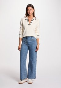 Frau steht mit einem weißen Langarmpullover mit schwarzer Besatz, hochgeschnittenen blauen Jeans und weißen Sneakers vor einem einfarbigen Hintergrund.