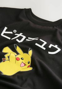 Must musta t-särk mustast puuvillast, millel on kollane Pikachu graafika ja jaapani keelne tekst. Krae on soonik, lisades kujundusele tekstuuri.