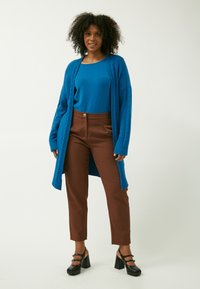 Cardigan blu in maglia con motivo a treccia, indossato sopra un top blu. Pantaloni marroni su misura con chiusura a bottone e scarpe con tacco a blocco nere.
