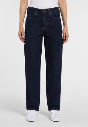 Osoba nosząca spodnie z ciemnoniebieskiego denimu o prostych nogawkach z przednimi kieszeniami oraz białe trampki, stojąca na tle jednolitego tła.