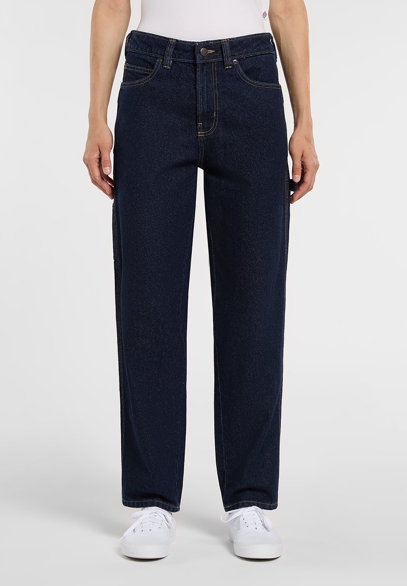 Person wearing straight-leg dark blue denim jeans with front pockets and white sneakers, standing against a plain background.