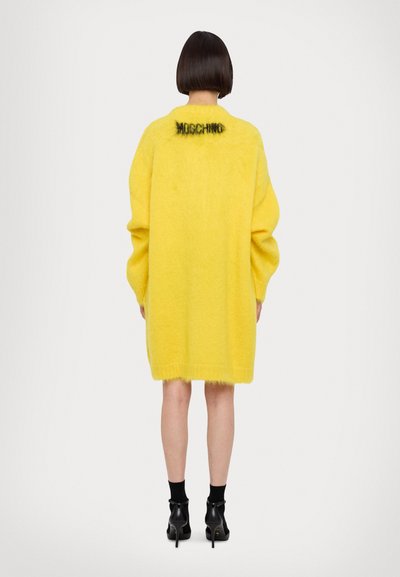Abito maglione oversize giallo in peluche con scollo rotondo, maniche lunghe e il nome del marchio lavorato a maglia in nero sulla parte superiore della schiena.