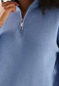 Eine Nahaufnahme einer Hand, die den Reißverschluss eines hellblauen, strukturierten Pullovers hält und das gerippte Muster des Stoffes hervorhebt.