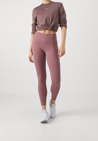 Femme debout dans des leggings mauves, un haut à manches longues noué assorti et des baskets blanches sur un fond clair uni.