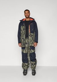 Veekindel rohelise ja mereväe toonides maastikuvärviga jope, millel on tõmblukud ja taskud, sobitatud sama mustriga pükstega. Komplekti lõpetavad mustad saapad.