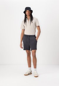 Beige kortærmet polo shirt med lynlås krave, parret med mørke teksturerede shorts og hvide sneakers med sorte accenter. Hatten er sort.