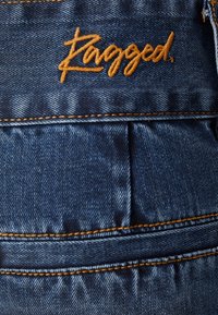Tummansininen denimi, jossa näkyvät saumat. Taskussa on ylhäällä vaakasuorien poimujen yläpuolella oranssiin ommeltu etiketti "Ragged.".